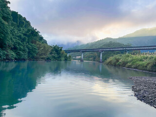 Serene early morning scene of Wulai River with clear turquoise water reflecting misty mountains and bridge in Wulai, New Taipei City, Taiwan. Golden sunlight breaking through clouds.