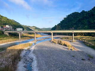 Early morning view of elevated bridge spanning wide rocky riverbed with shallow stream in Wulai, New Taipei City, Taiwan. Golden light on lush green mountains.
