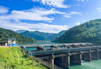 The industrial structure of Zhihtan Weir Dam, or Zhihtan Dam, controls the turquoise water flow in Xindian, New Taipei City, Taiwan. Lush forested mountains rise under a bright blue sky.