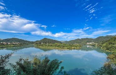 Wide view of Qingtan Weir, Xindian, New Taipei City, Taiwan. Turquoise water perfectly mirrors the blue sky and clouds. Lush green hills frame the tranquil scene in the afternoon.