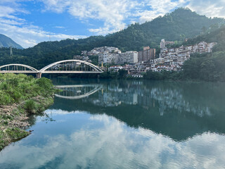 The tranquil waters of Swallow Lake, Xindian, New Taipei City, Taiwan, reflect the sky and surrounding green mountains. An elegant arch bridge connects a charming riverside town.