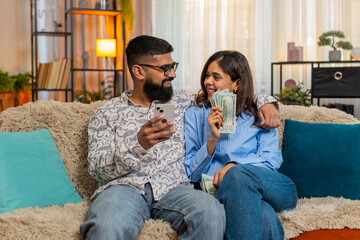 Indian couple man and woman sitting on home sofa holding money dollar cash and smartphone while planning purchases and family budget. Discussing future expenses, savings and income from salary indoors