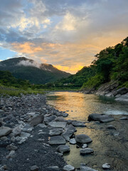 Serene dawn landscape in Wulai, New Taipei City, Taiwan. Golden sky reflects on peaceful river surrounded by mountains, forests, and smooth rocks along shore.