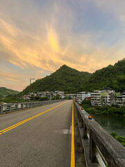 Empty bridge leading into Wulai village under a dramatic orange sunrise sky with wispy clouds, surrounded by lush green mountains, New Taipei City, Taiwan.