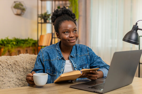 African American young woman sitting at table on home sofa reads an engaging book with deep concentration, calmly turning pages. Black girl enjoys the quiet moment of focused relaxation and interest.