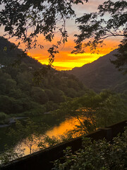 A blazing orange and yellow sunrise reflected in the river water, framed by dark tree branches and mountains in Wulai, New Taipei City, Taiwan, capturing natural intensity.