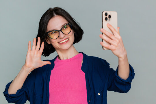 Young woman blogger taking portrait selfie on smartphone, communicating record video call online with social media subscribers. Girl demonstrates enthusiasm lively communication on gray background