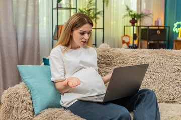 Pregnant middle-aged woman at home sees laptop freeze waves hand upset sets device aside feels annoyed loudly. Caucasian girl on sofa struggles with tech error seeks quick fix before baby time soon.