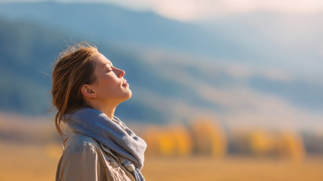 A woman stands outdoors with her eyes closed savoring the peacefulness of nature. The soft sunlight illuminates the landscape highlighting her calm demeanor and the distant mountains.
