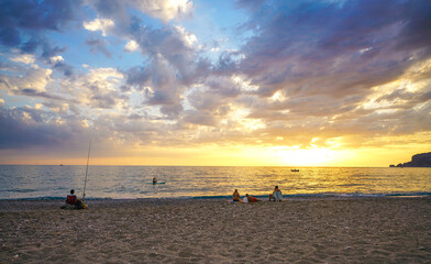 A warm, beautiful sunset over the sea and a pebble beach, where several people are relaxing, fishing, and enjoying the twilight. A tranquil coastal nature scene with a dramatic sky and golden light.