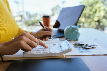 Obraz premium A person in a yellow sweater places a coin into a blue piggy bank on a desk with charts, coins, a laptop, and a coffee cup, symbolizing saving and financial planning.