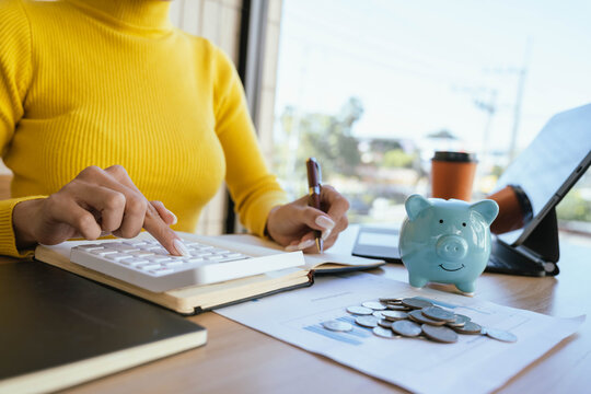 A person in a yellow sweater places a coin into a blue piggy bank on a desk with charts, coins, a laptop, and a coffee cup, symbolizing saving and financial planning.