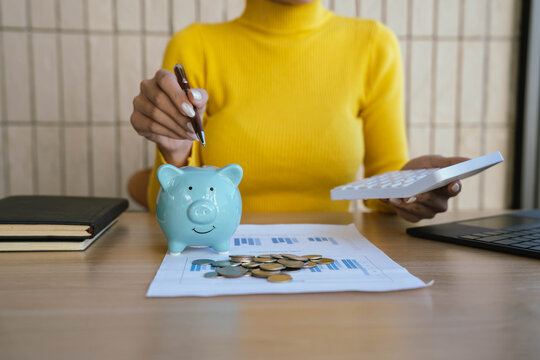 A person in a yellow sweater places a coin into a blue piggy bank on a desk with charts, coins, a laptop, and a coffee cup, symbolizing saving and financial planning. - Powered by Adobe
