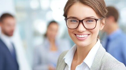 A confident woman wearing glasses smiles warmly in a bright office. Colleagues can be seen discussing in the background showcasing a collaborative work environment.