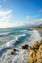 Fototapeta premium Vertical view of dramatic turquoise waves crashing onto a rocky coast, leading to a sunny resort town and mountains in the distance. Vibrant seascape, travel, and vacation concept.
