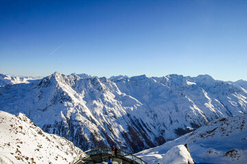 Naklejka premium A stunning panoramic view of the snow-capped Alps under a clear blue sky. A winter landscape with majestic peaks, icy slopes, and a glass observation deck overlooking the valley.