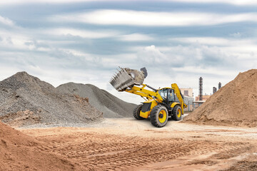 A yellow loader operates on a construction site, lifting sand and gravel with piles of materials...