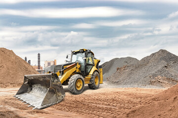 A yellow backhoe loader moves dirt and gravel at a construction site. Heavy clouds fill the sky,...