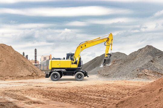 A yellow excavator operates on a construction site, digging and moving material next to large mounds of sand and gravel under an overcast sky