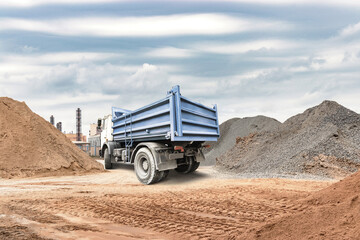 A truck is unloading materials at a construction site filled with piles of sand and gravel under a cloudy sky in a busy industrial area © Anoo
