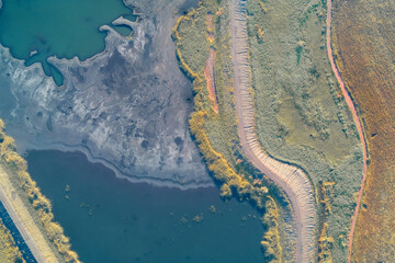 Top-down drone view capturing the sharp boundary created by a winding dirt road between a dark contaminated lake and a tilled brown field. Severity of the ecological crisis and environmental damage