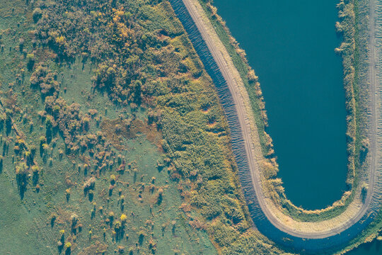 Abstract drone perspective capturing deep blue-green contaminated water body tightly contained by curved embankment. Wild lush vegetation highlights the ecological crisis and human control over nature