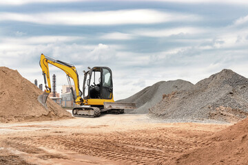 An excavator digs at a construction site surrounded by large mounds of sand and gravel. The sky is...