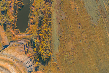 Abstract drone sharp boundary between a terraced industrial quarry and an agricultural field. The autumn trees and a small pond highlight the contrast between nature, mining, and human land use