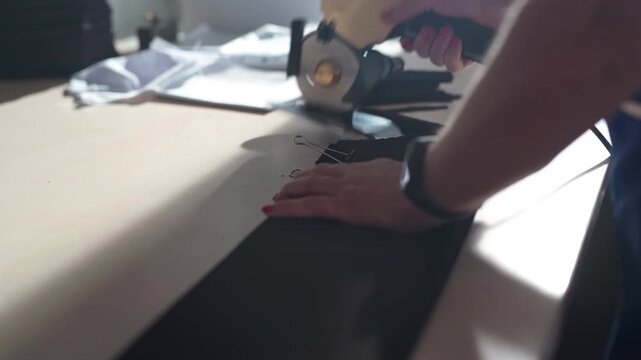 A close view shows a rotary fabric cutter slicing stacked black technical material while a hand steadies the layers on the work table under soft daylight