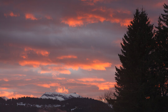 Fiery red clouds glow above snow-topped alpine peaks and dark forest silhouettes during a dramatic winter sunset in the mountains.