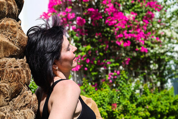 A young woman stands with her back to a palm tree against wall of flowers.