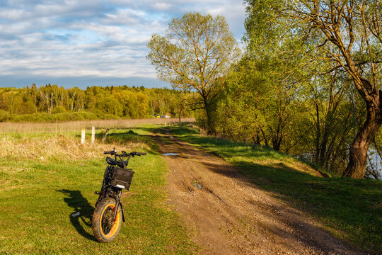 Rugged fat tire electric bike parked on grassy verge next to a dirt track winding through bright spring countryside at sunset
