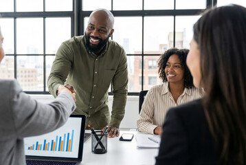 Smiling Black businessman shaking hands with partner during diverse team meeting in modern office with data charts
