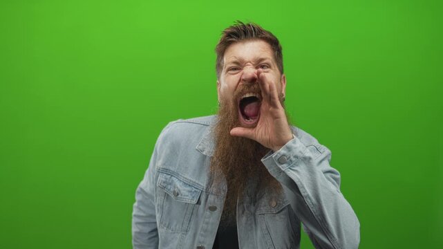 Man with long beard wearing denim jacket and hands cupping mouth shouting in green studio; urgent announcement.