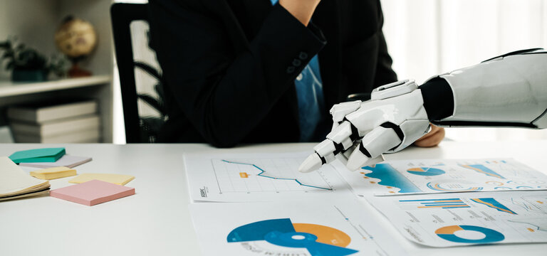 A business professional engages in data analysis while a robotic arm highlights financial charts, showcasing the intersection of technology and corporate strategy in the modern workplace.Noogenesis - Powered by Adobe