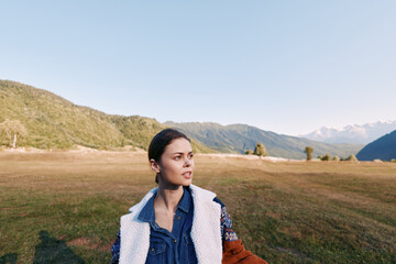 Fototapeta premium Woman portrait in meadow with mountains landscape, outdoor nature and travel mood. Young female in denim jacket and shearling coat gazing to the side across grassy field.