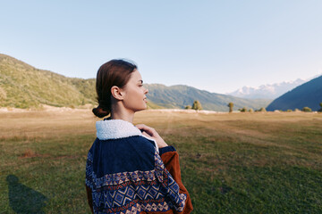 Fototapeta premium Woman in patterned jacket standing in meadow, portrait and profile view with mountains and landscape in background. Travel scene with serene nature, grass field and clear sky.