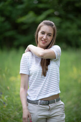 Fototapeta premium Young woman with long brown hair, wearing a striped shirt and light pants, stands in a lush green field, smiling warmly, embodying a sense of joy and connection with nature