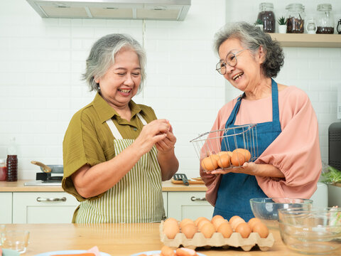 Happy senior Asian women preparing to cook with fresh eggs in a kitchen. Cheerful elderly friends holding a basket of eggs and laughing together.