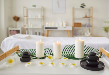 Serene spa setting with burning candles, black stones and white flowers arranged on table. Modern interior of massage or cosmetology room with relaxing decor and massage table on background.