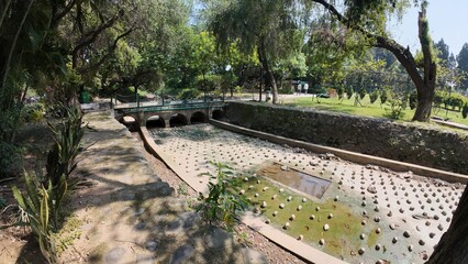 Small Arched Stone Bridge Spanning a Dry, Stone-Lined Water Canal or Channel In a City Park or Recreational Area on a Bright, Sunny Day