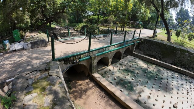 Small Arched Stone Bridge Spanning a Dry, Stone-Lined Water Canal or Channel In a City Park or Recreational Area on a Bright, Sunny Day - Powered by Adobe