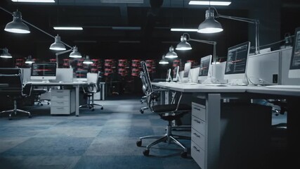 Empty office space with rows of computers and desks illuminated by overhead lamps in a dark modern workspace showing data visualizations on screens - Powered by Adobe