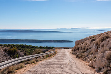 Fototapeta premium A steep concrete road descends through rocky terrain towards the Adriatic coast. This scenic route on Kamenjak mountain offers breathtaking panoramic views of the nearby islands and sea in Croatia.