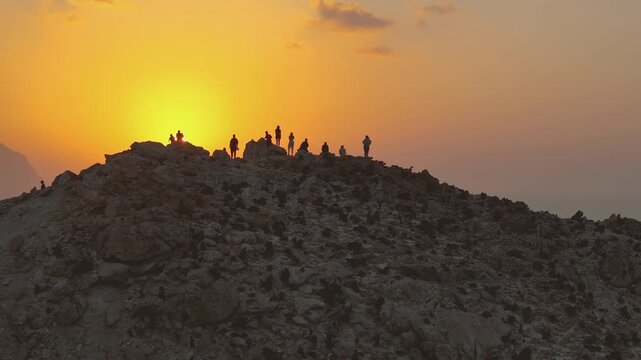 A group of people stand on a rocky mountain ridge, silhouetted against a vivid orange sunset, with rugged terrain and distant peaks fading softly into the warm evening light.