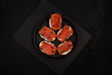 Salted red fish sandwiches on bread with cream cheese and radish microgreens on a black plate on a stone cutting board against a black background. Top view with copy space. Healthy eating food photo