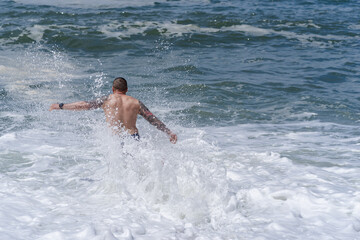 Back view of a tattooed man standing amidst rough white foam waves of the Atlantic Ocean on a sunny day.