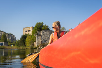 Blonde woman paddling a red canoe past the submerged prison ruins at Rummu Quarry, Estonia on a sunny summer day.