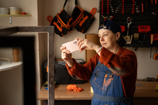 Young professional mechanic or craftsman woman in blue overalls take photo on mobile phone fix work result in repair renovation workshop.