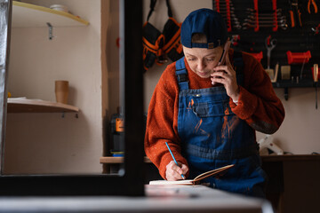 Craftsperson making notes in notepad to make orders from clients on smartphone. Manufacturer preparing restoring project in workshop